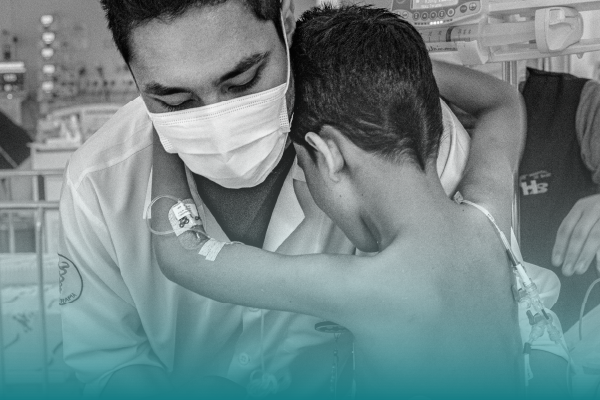 Foto em preto e branco de um menino de aproximadamente dez anos com as mãos em volta do pescoço de um fisioterapeuta que usa máscara cirúrgica e jaleco. O garoto está sem camisa e de costas para nós. Ambos são vistos da barriga para cima. Ao fundo, leitos hospitalares.